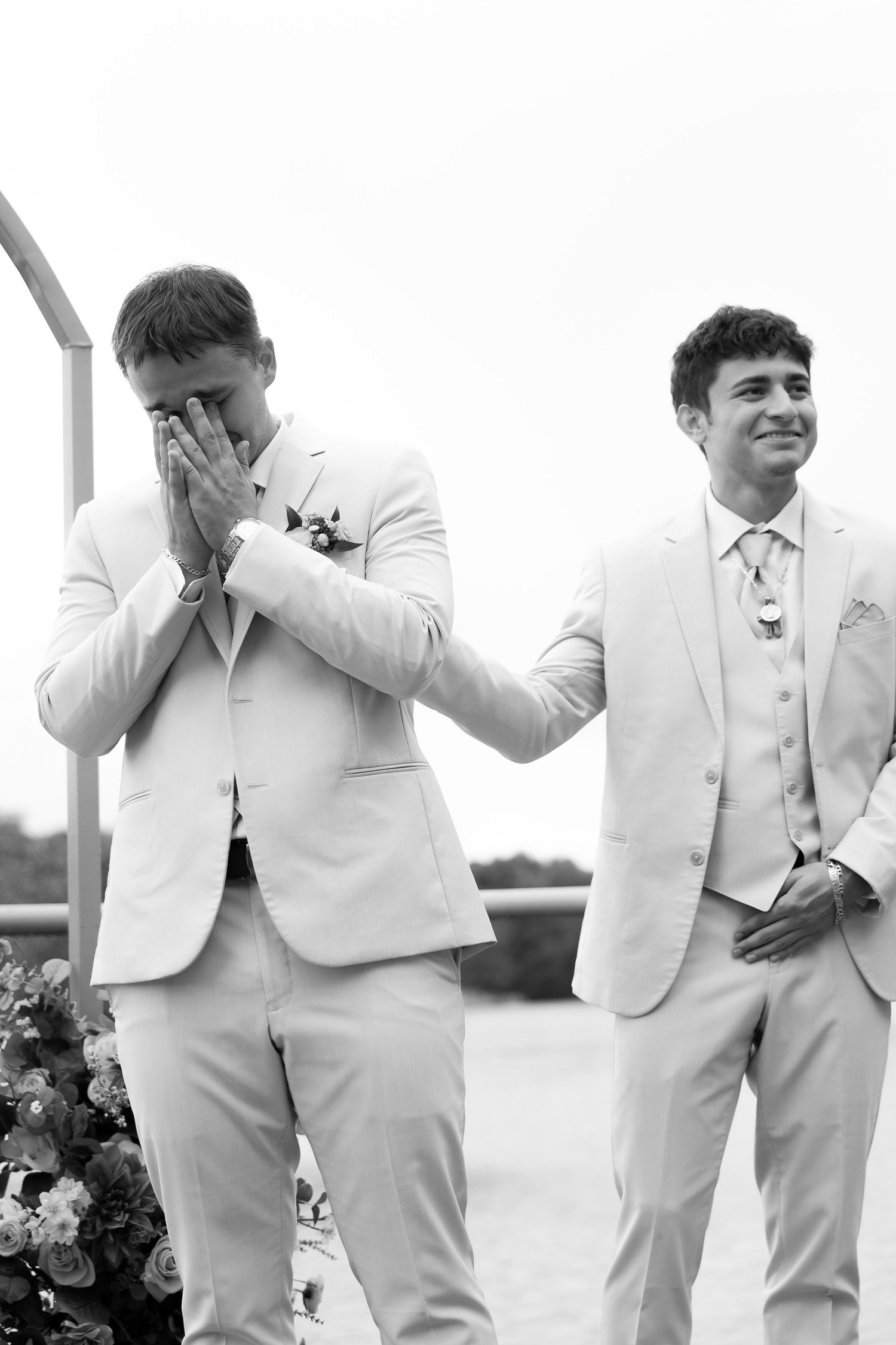Two groomsmen in cream suits at wedding altar, one emotional covering face, other smiling supportively.