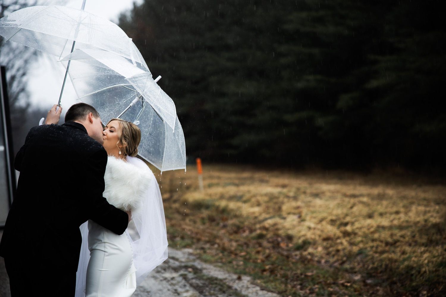 A romantic rainy day wedding scene with a clear umbrella against a moody background.