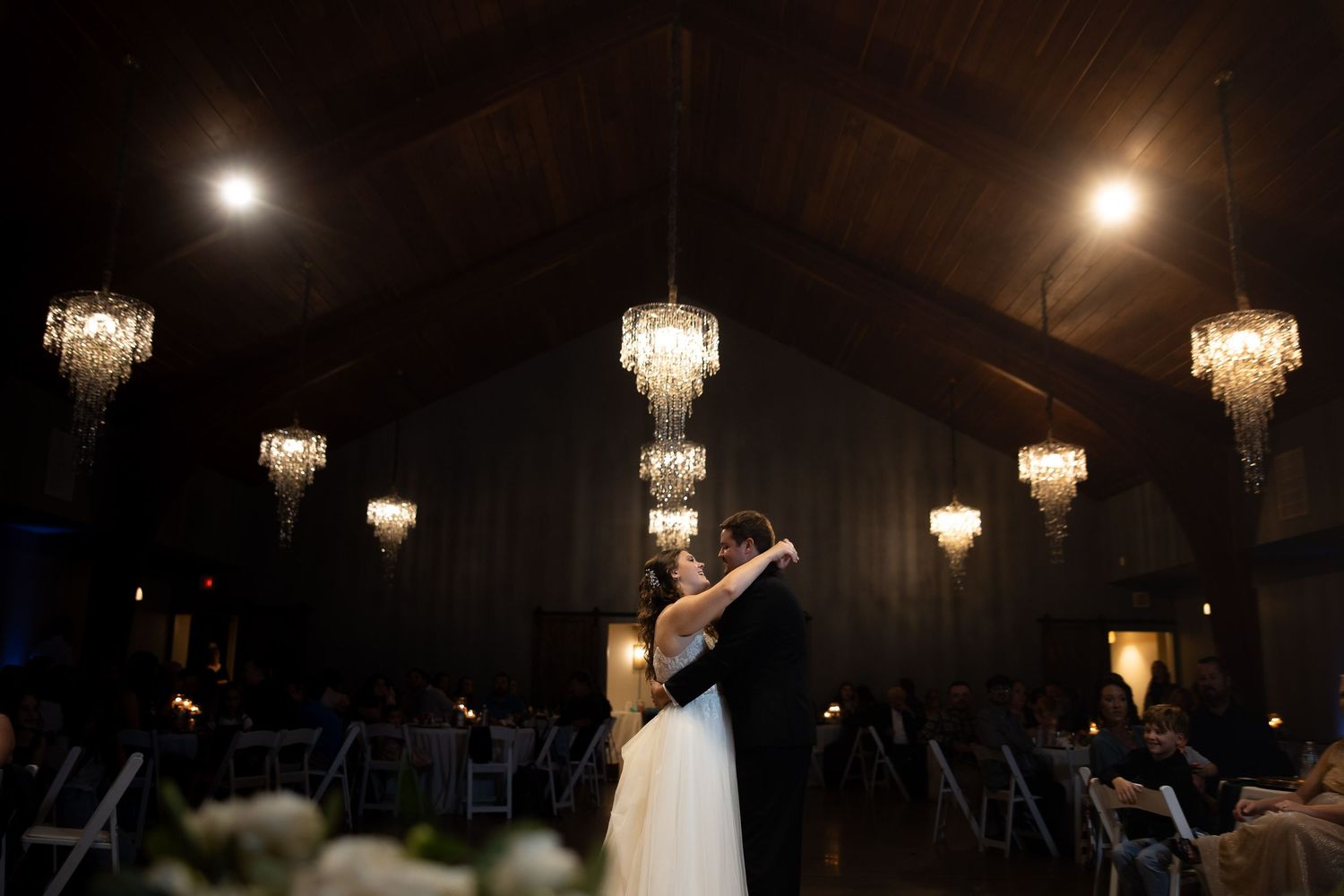 Bride and groom share first dance under crystal chandeliers in elegant dark reception hall.