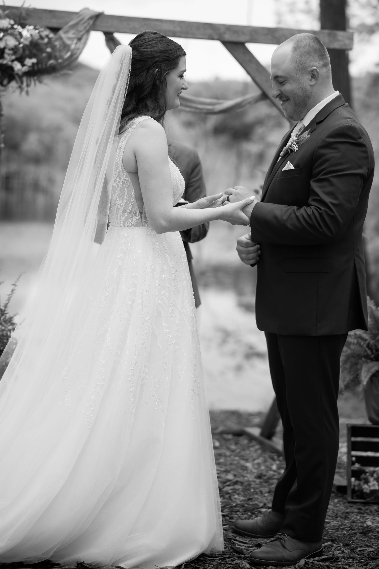 Black and white photo of bride placing wedding ring on groom's finger during outdoor ceremony.
