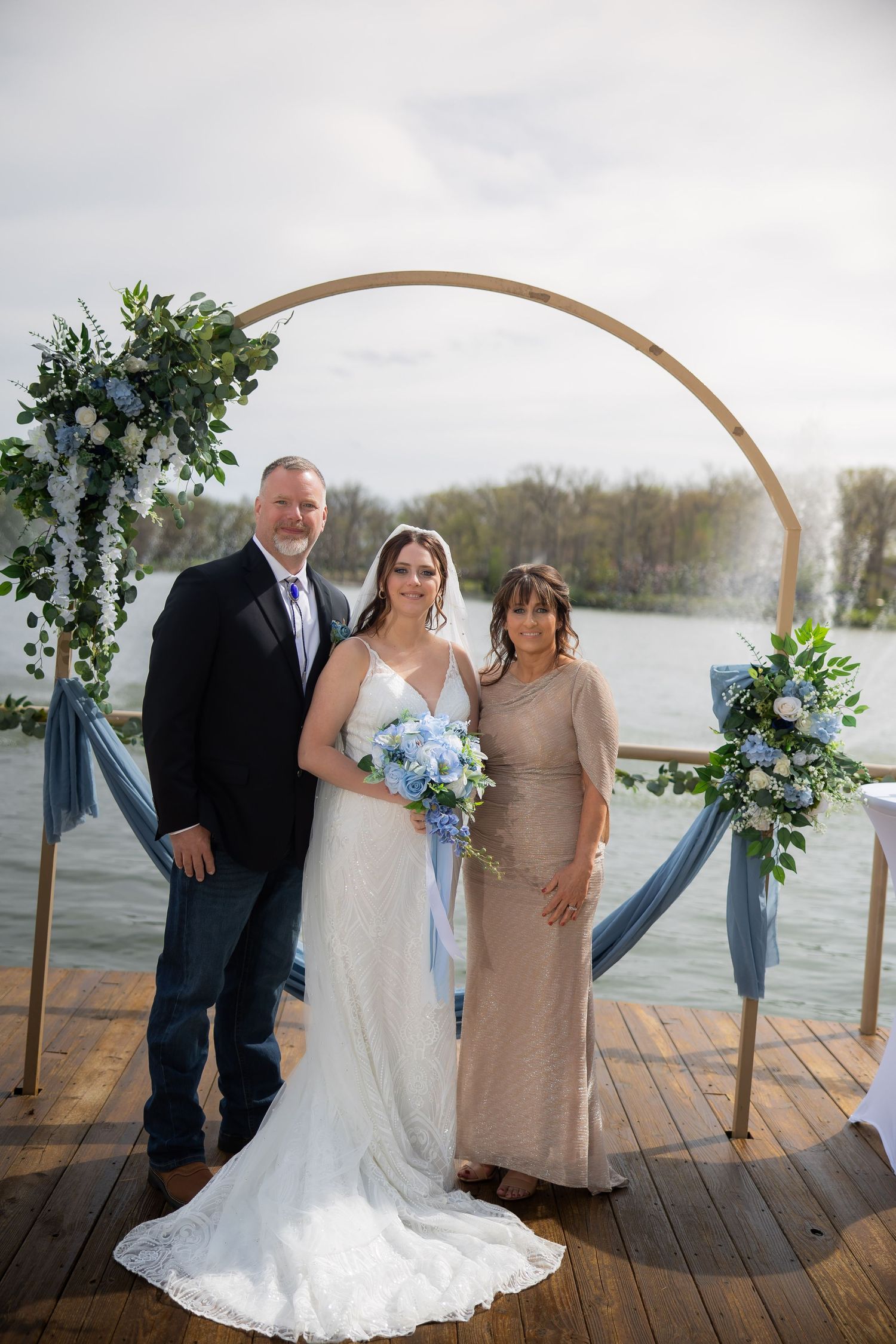 Bride in white gown holds blue bouquet with two guests under floral arch on waterfront dock at outdoor wedding ceremony.