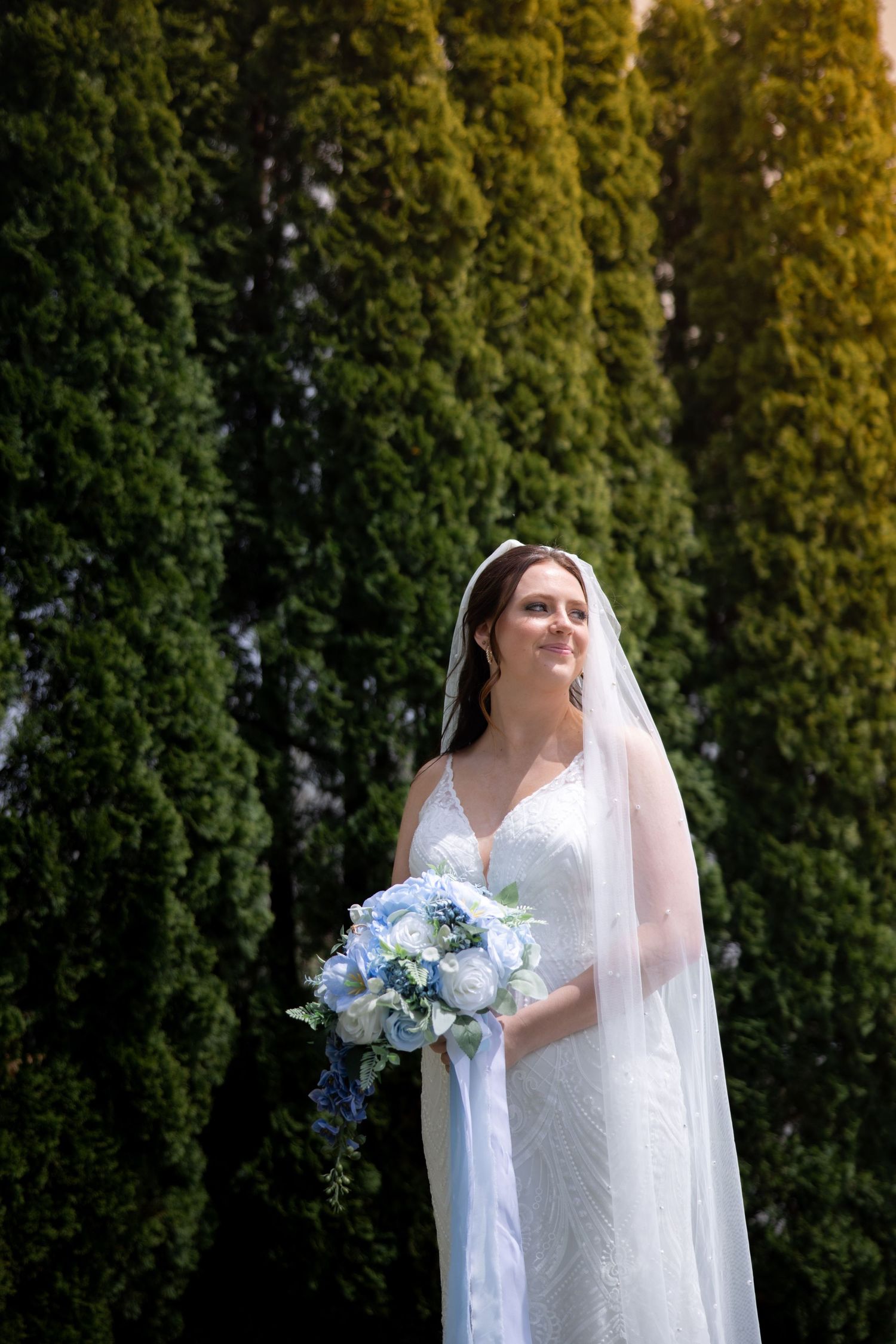 Bride in white lace gown and veil holds blue and white bouquet, standing before tall green cypress trees outdoors.