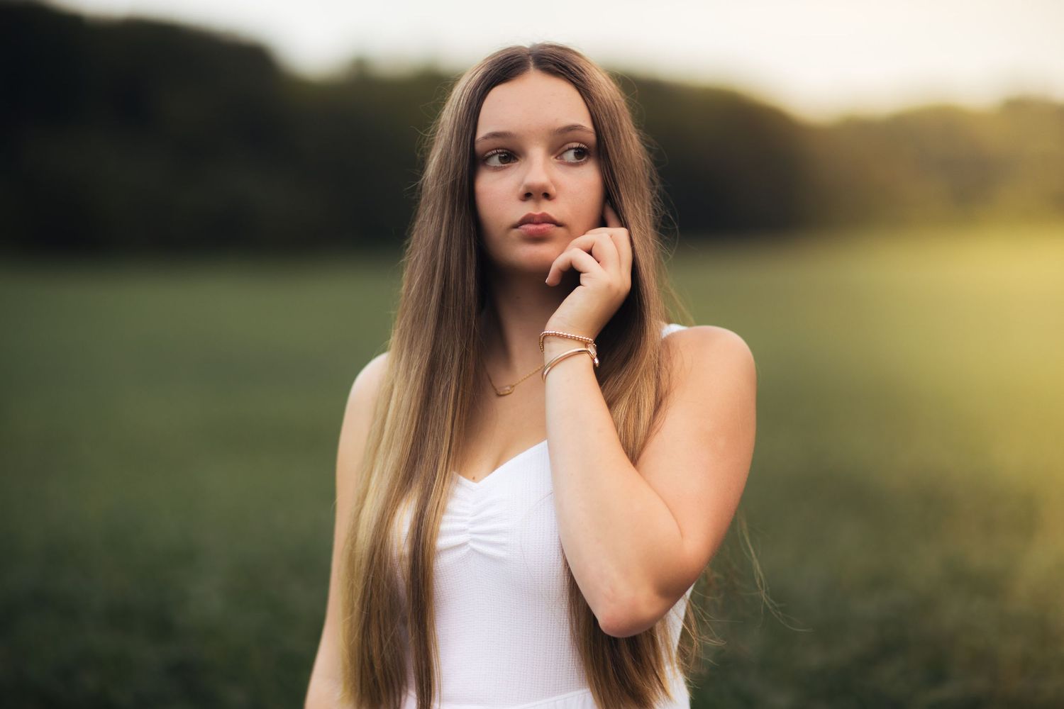 A person with long straight hair poses in a white top against a sunset-lit field.