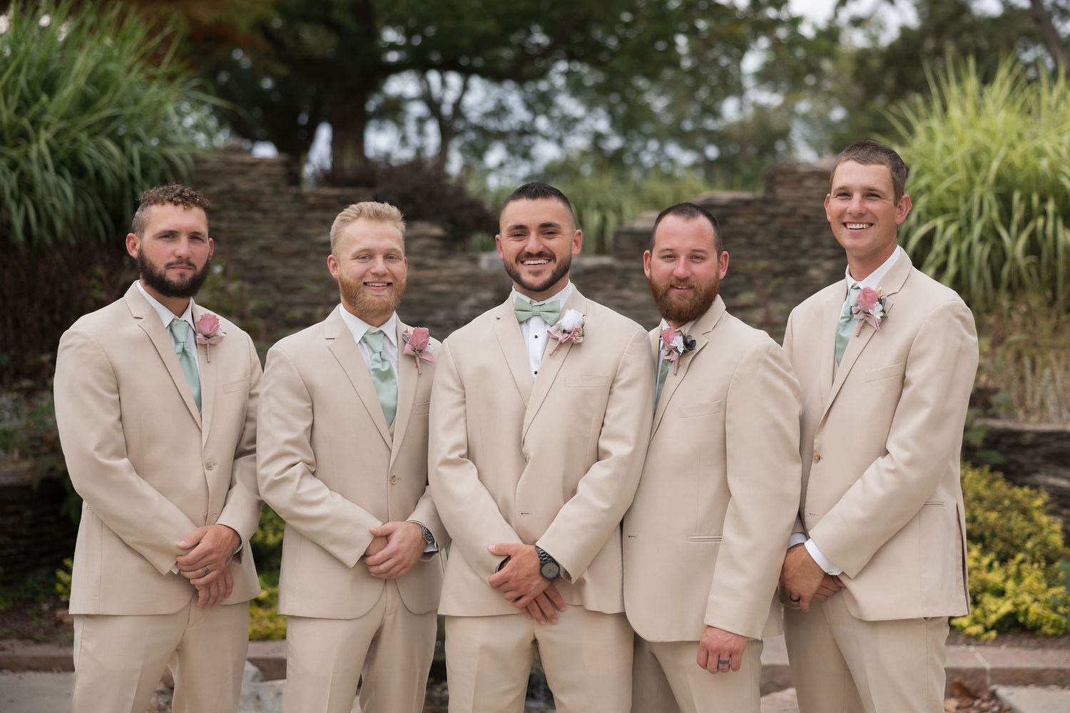 Groomsmen in tan suits pose together outdoors for formal wedding party photos.