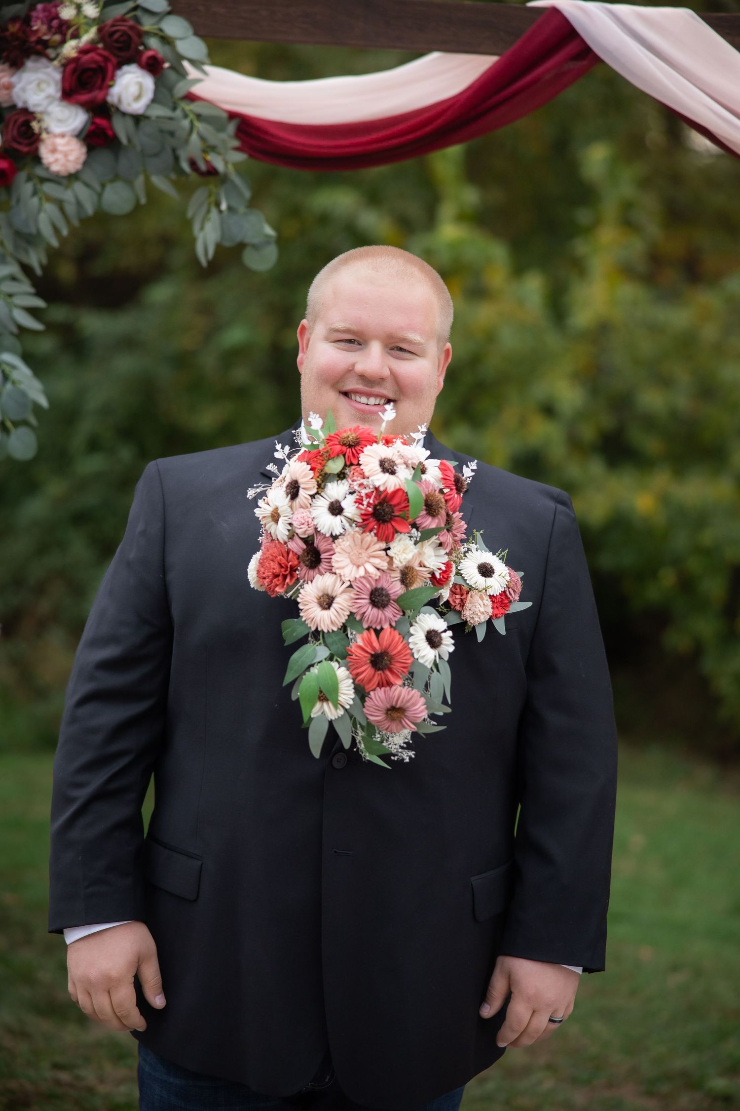 A person in a black suit stands under a wedding arch decorated with red and pink floral arrangements at an outdoor ceremony.