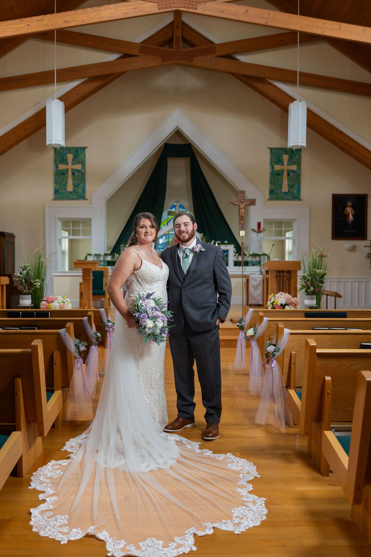 Bride and groom pose together in a charming church aisle, bride wearing lace gown with long train holding purple bouquet.