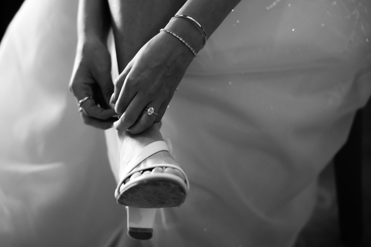 Bride adjusting her white wedding shoe, showcasing an elegant diamond ring and delicate bracelet in black and white.