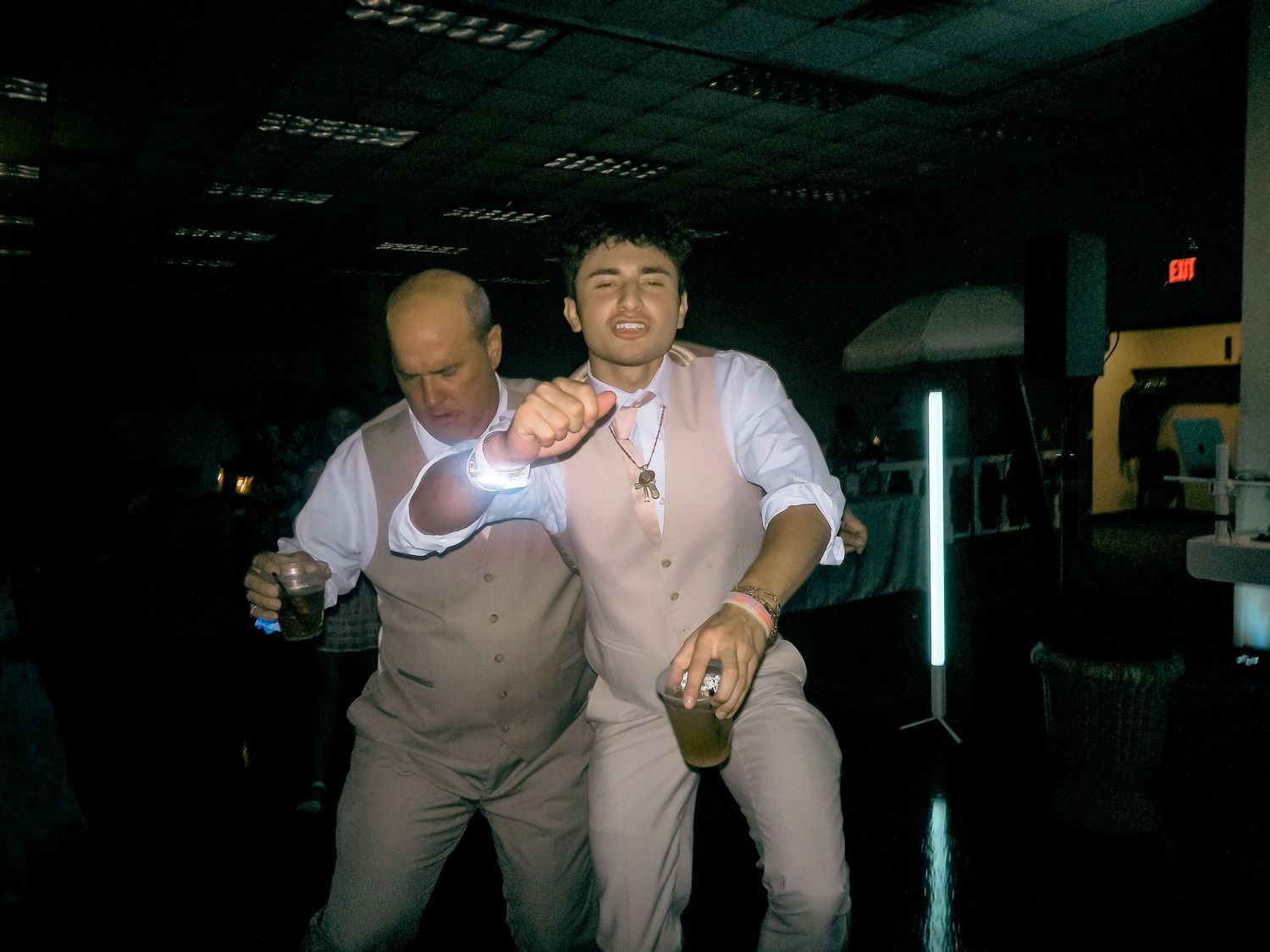 Two men in white suits dancing and holding drinks at a dimly lit party or wedding reception.