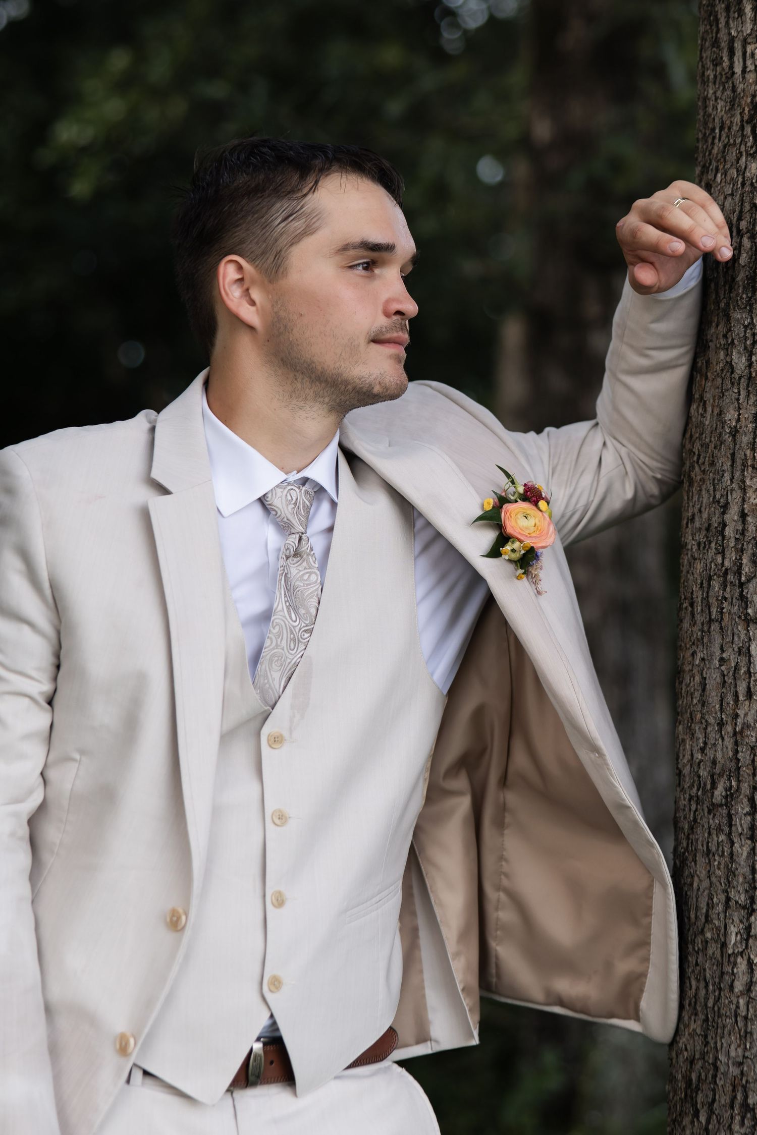 Groom in beige suit with boutonniere leaning against tree in outdoor portrait.