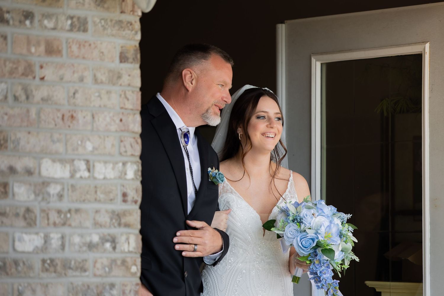 Bride in white gown holding blue bouquet stands with older man in suit near brick wall before wedding ceremony.