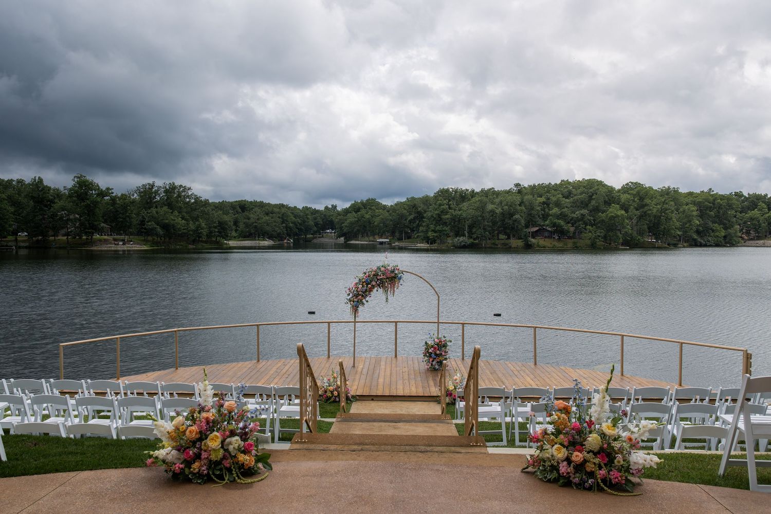 Outdoor lakeside wedding ceremony setup with white chairs, floral arch, and dramatic cloudy sky over serene water.