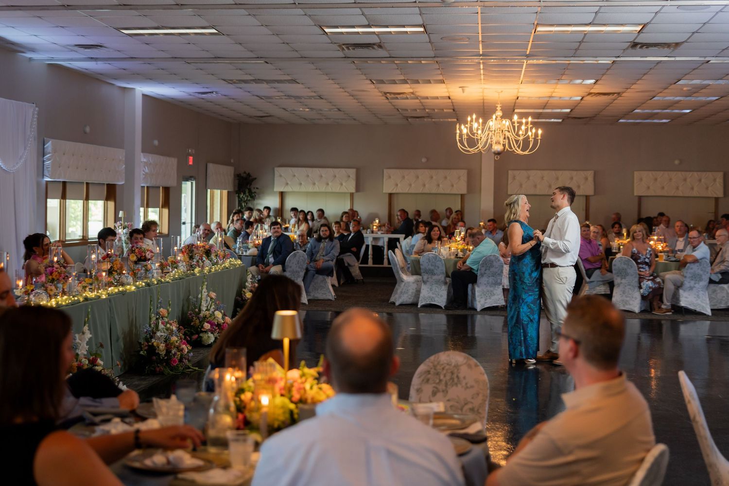 Wide view of elegant wedding reception hall with chandelier, candles, and couple dancing for guests.
