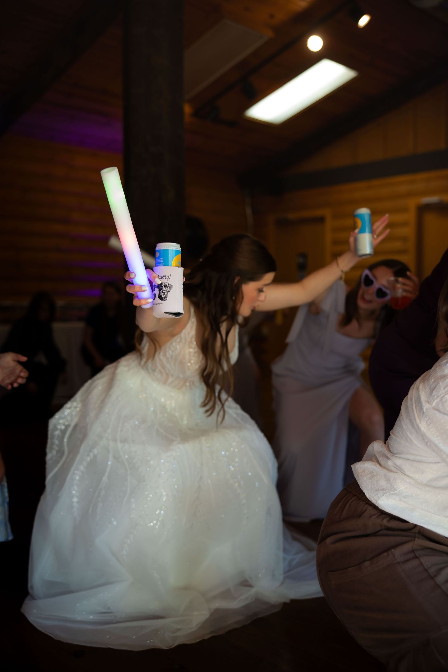 Bride in white gown dances with a glow stick and drink at a lively rustic wedding reception.