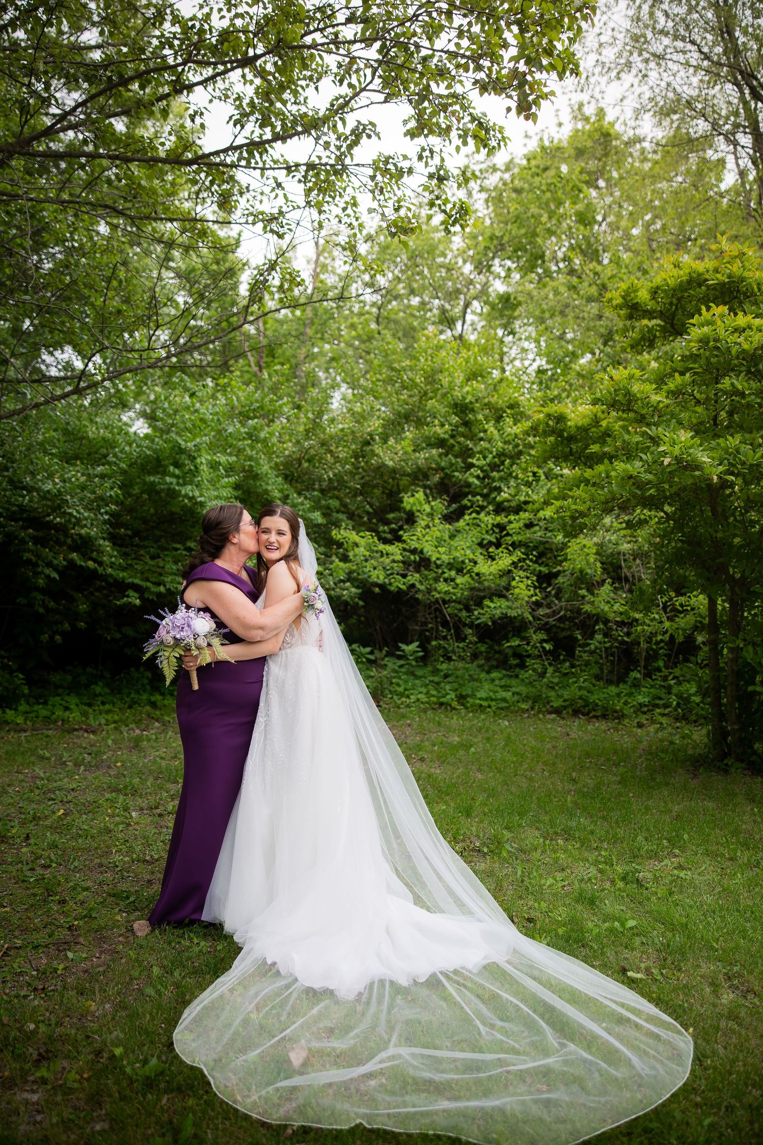 Bride in long white gown with cathedral veil embraces woman in purple dress in a lush green outdoor setting.
