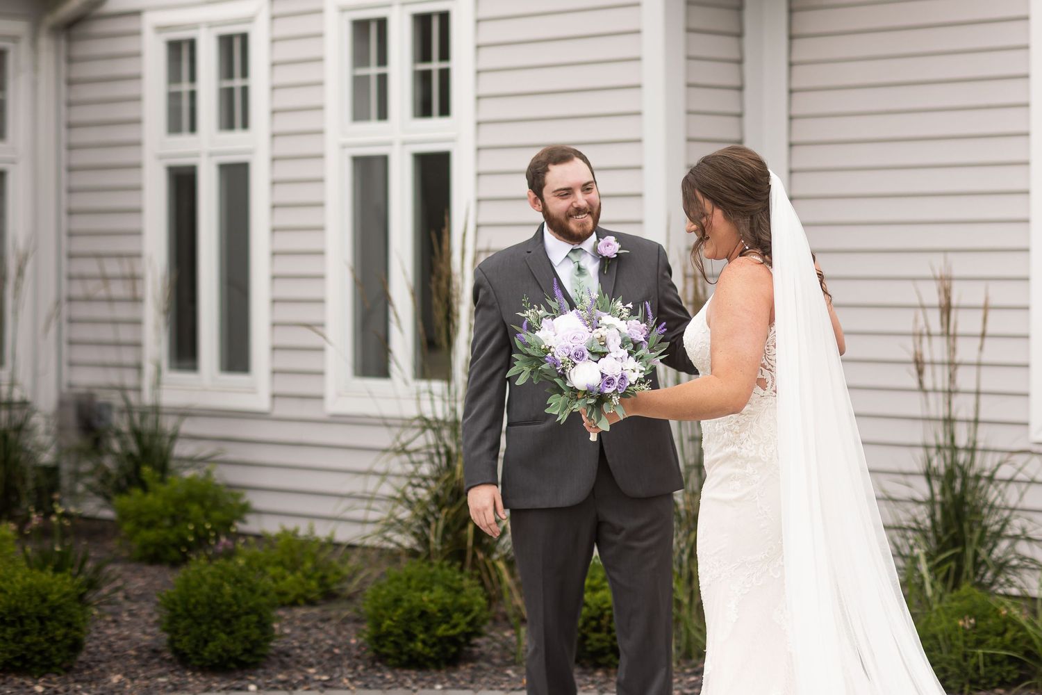 Groom smiles at bride holding a lavender bouquet during first look outside gray building.