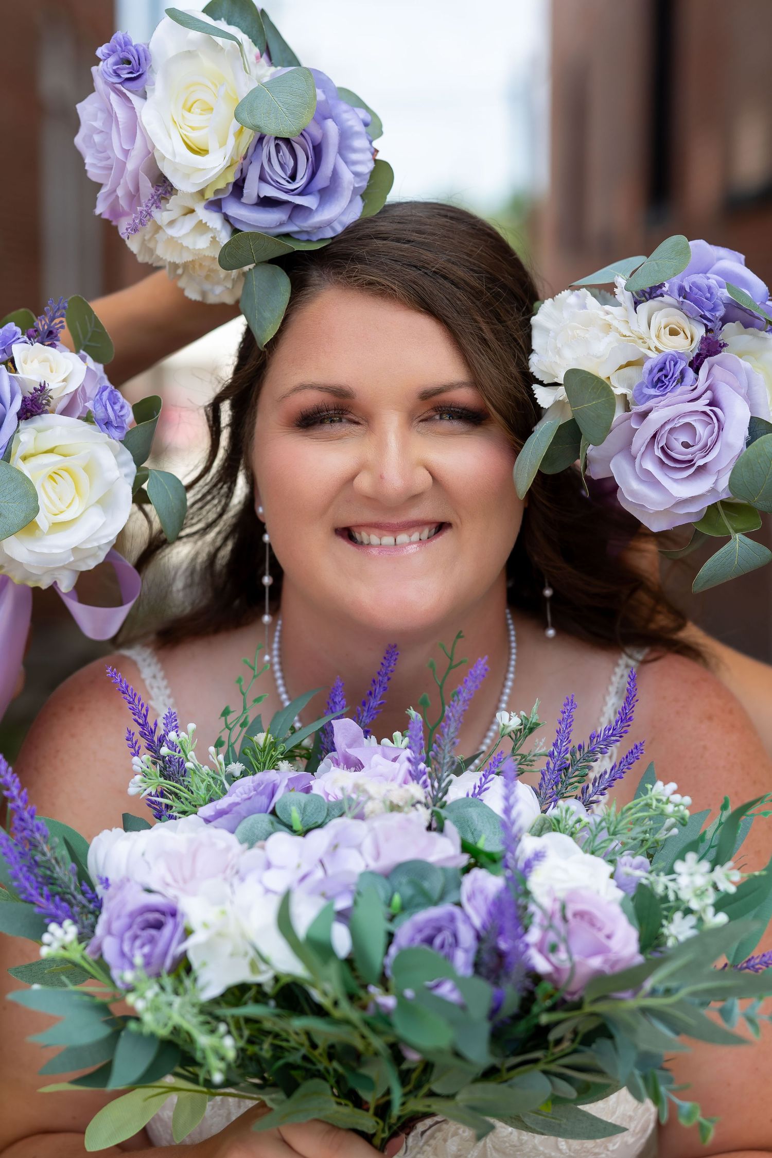 Smiling bride surrounded by purple and white rose bouquets on her wedding day.