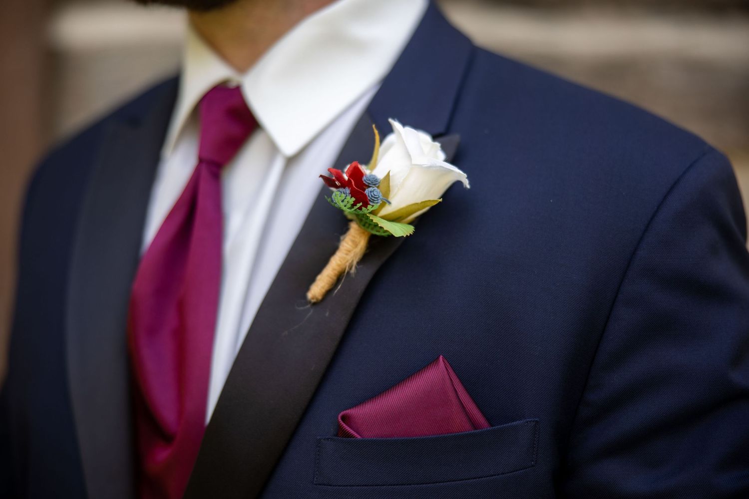 Groom in navy suit with burgundy tie, white rose boutonniere, and matching pocket square for a wedding.