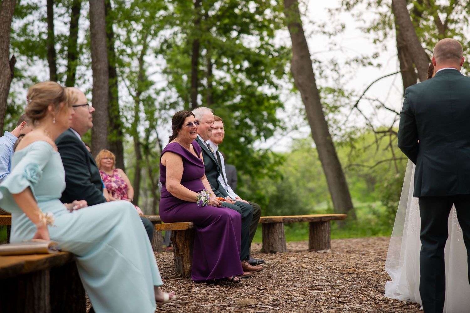 Wedding guests seated on rustic wooden benches during an outdoor forest ceremony celebration.