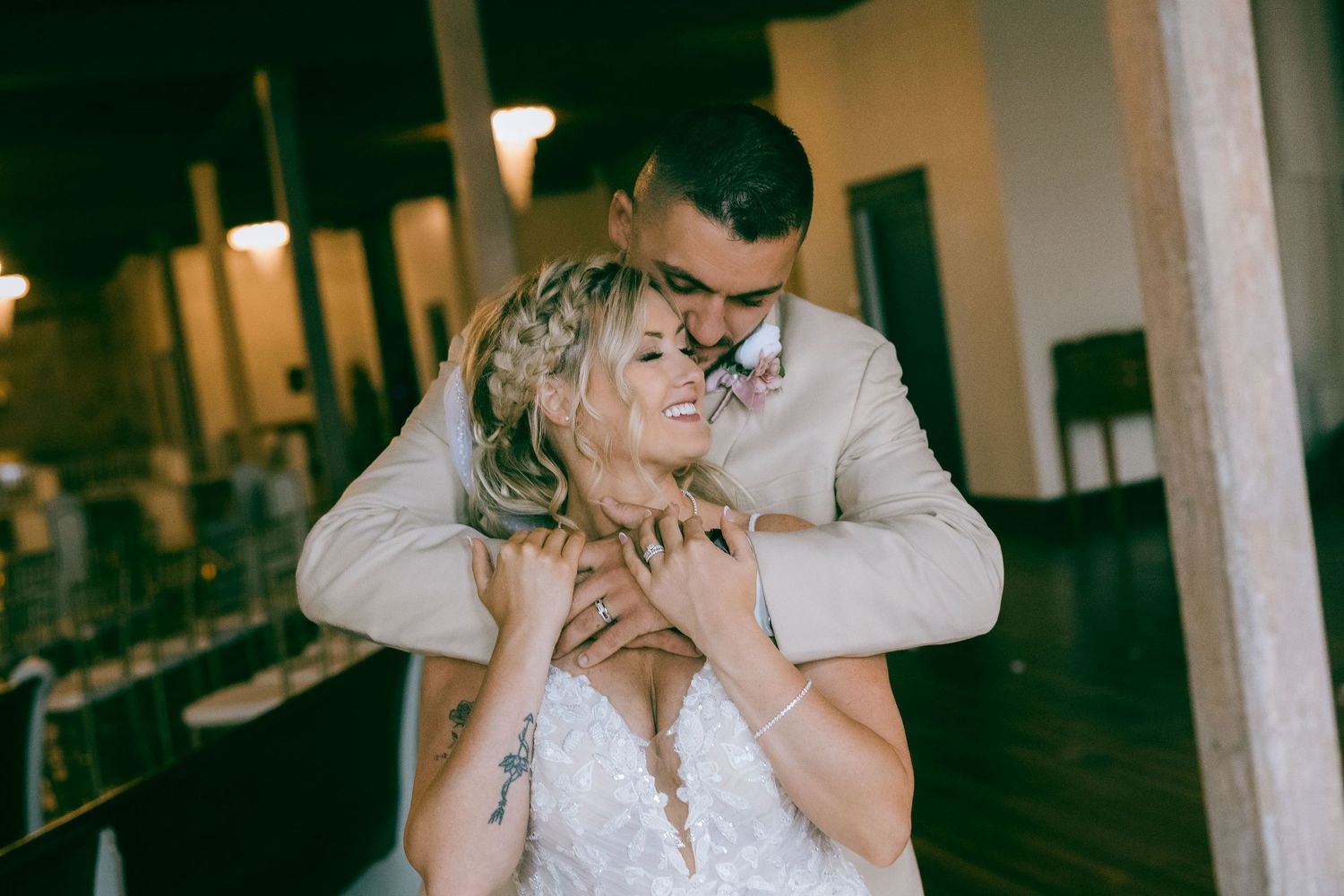 Wedding couple embraces in rustic indoor venue with natural lighting and romantic atmosphere.