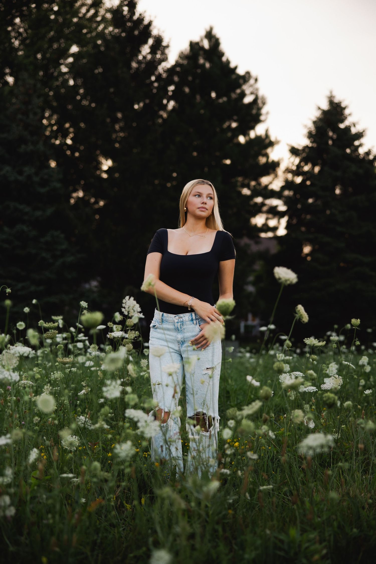 Person wearing black top and distressed jeans stands among blooming white wildflowers at dusk.