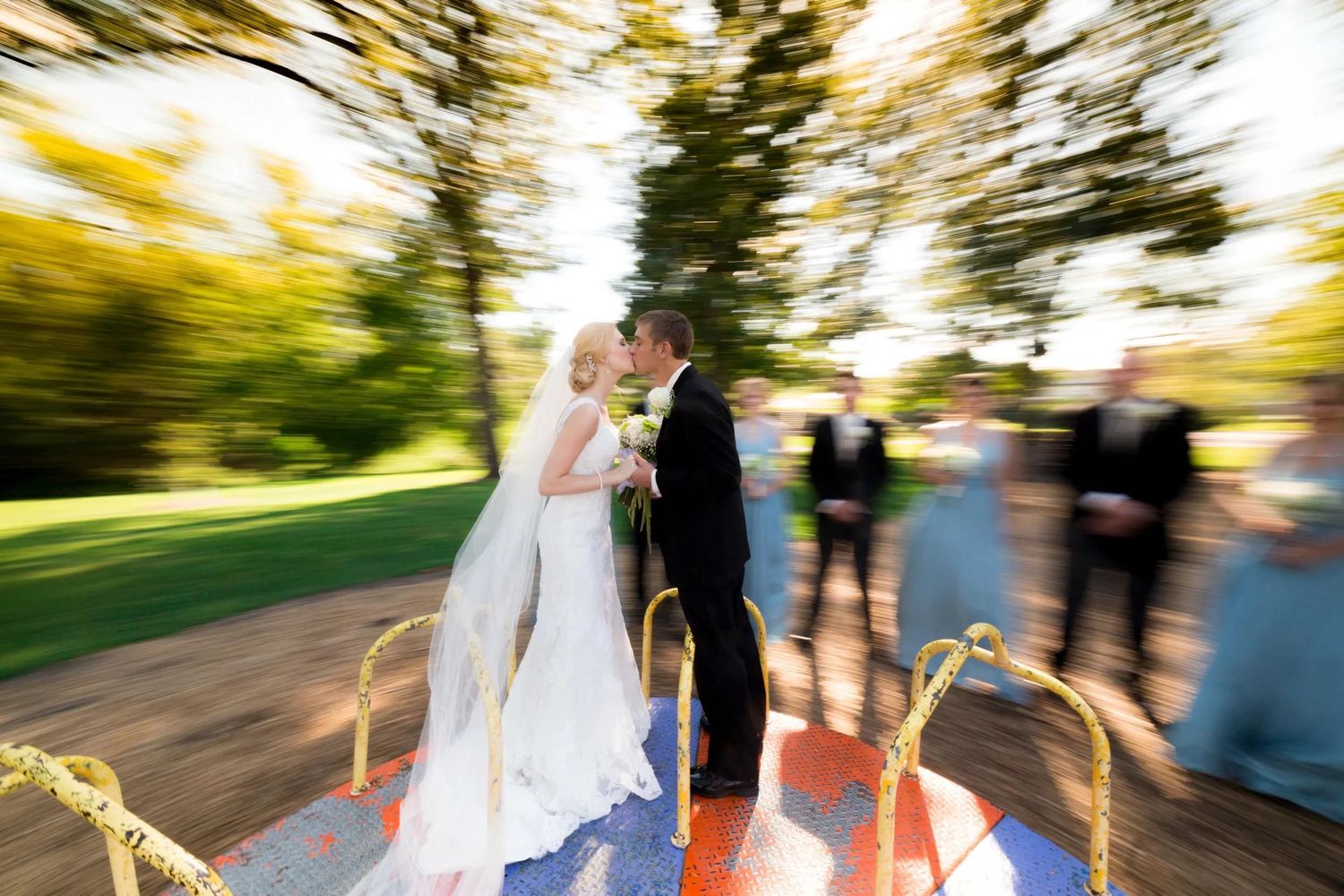 Bride and groom share a kiss on a carousel at their outdoor wedding ceremony, with guests visible in the background.