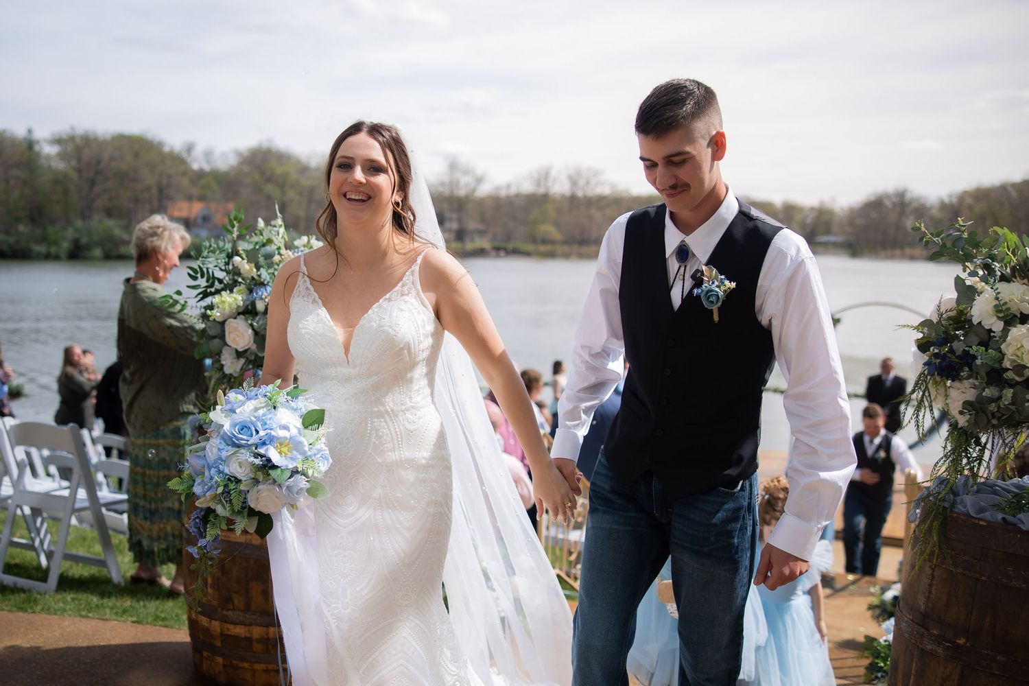 Smiling bride and groom walk hand-in-hand after outdoor waterfront wedding ceremony, bride holding blue and white bouquet.