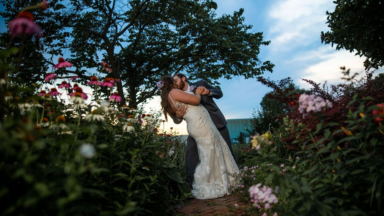 Bride and groom share romantic embrace in lush garden surrounded by colorful flowers under a twilight sky.
