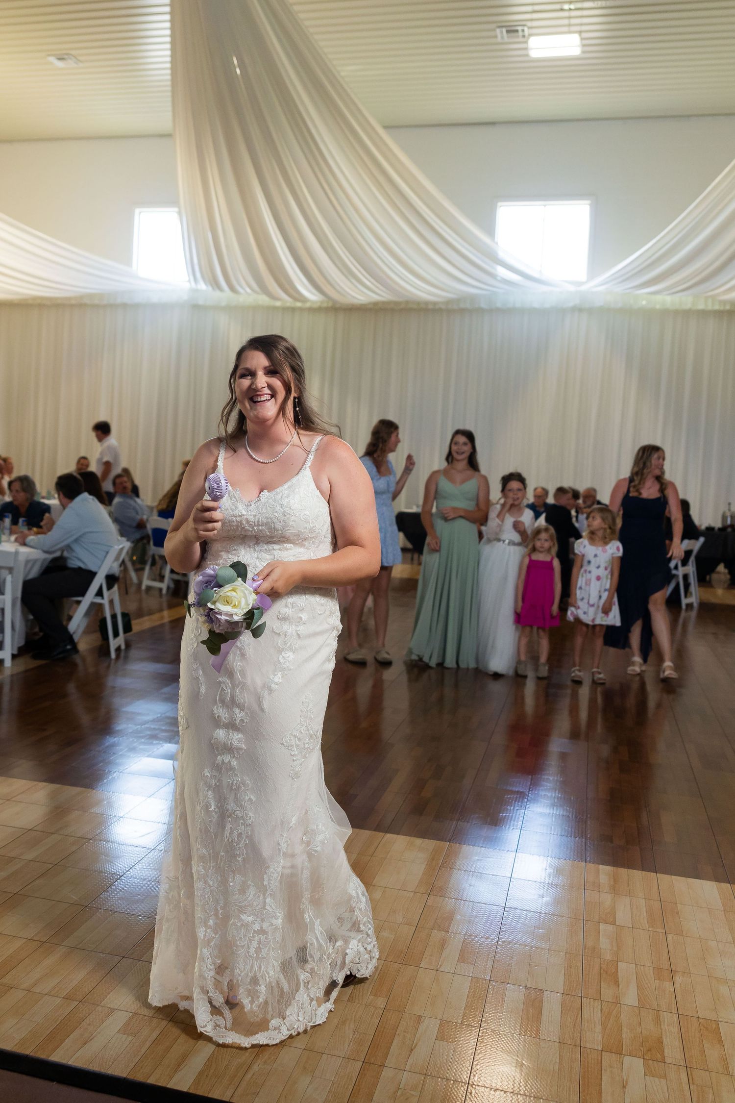 Smiling bride in lace wedding gown holding bouquet on dance floor at elegant indoor wedding reception.