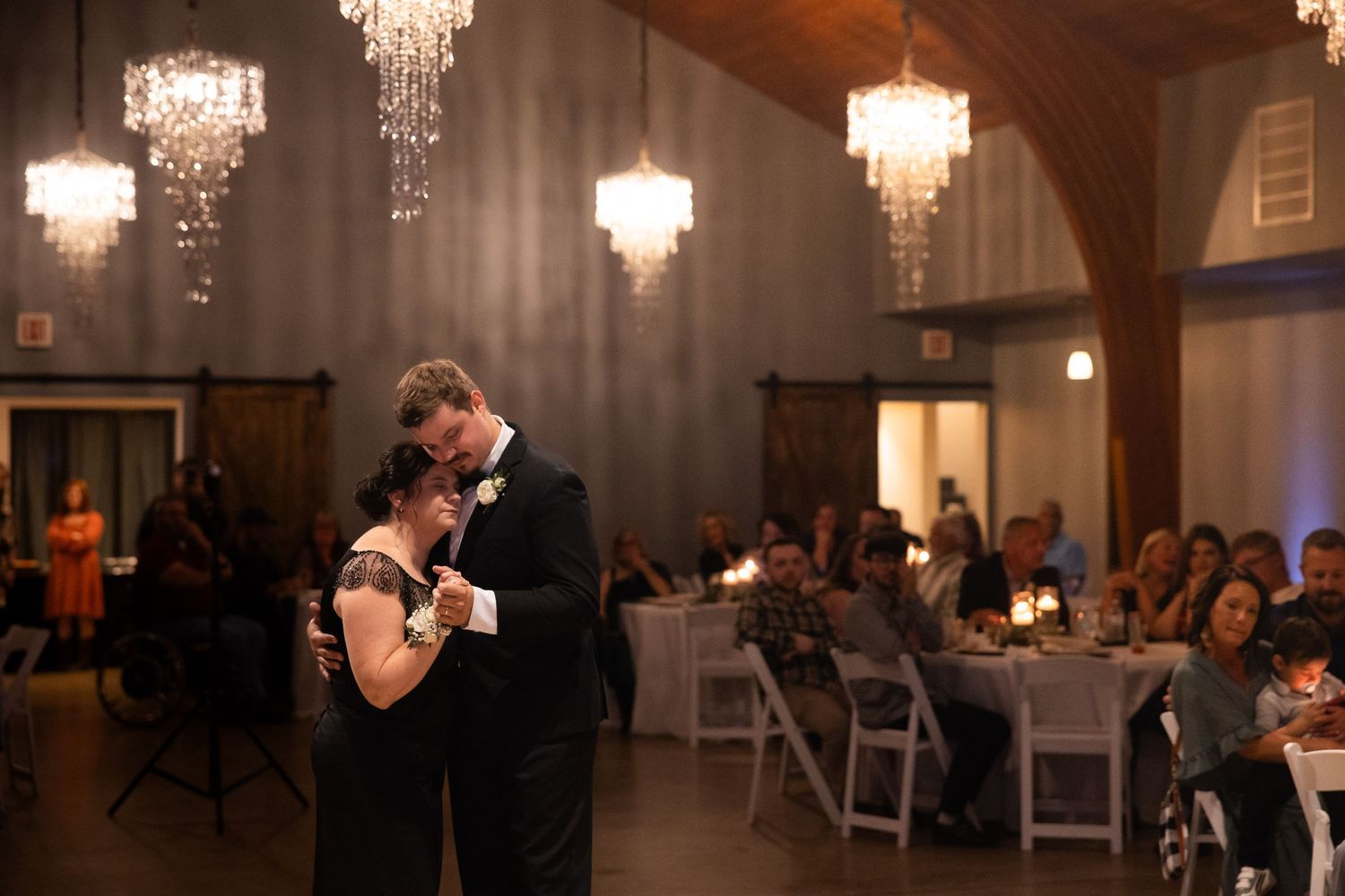 Couple shares first dance at elegant wedding reception with chandeliers and guests seated at candlelit tables.