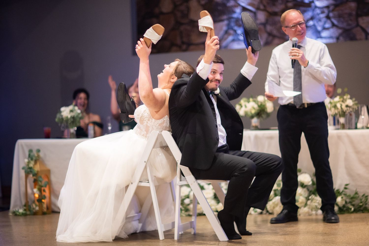 Bride and groom laughing and raising shoes during wedding reception game while officiant holds microphone nearby.