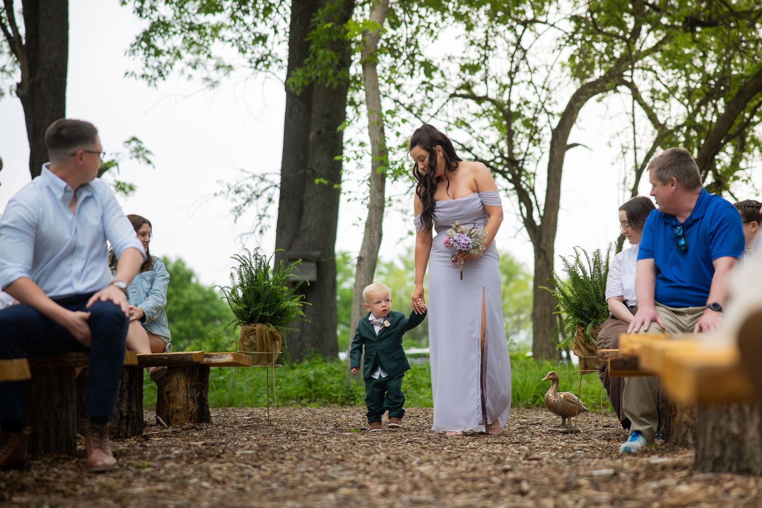 Bridesmaid in lavender gown walks young ring bearer in suit down wooded outdoor wedding ceremony aisle.