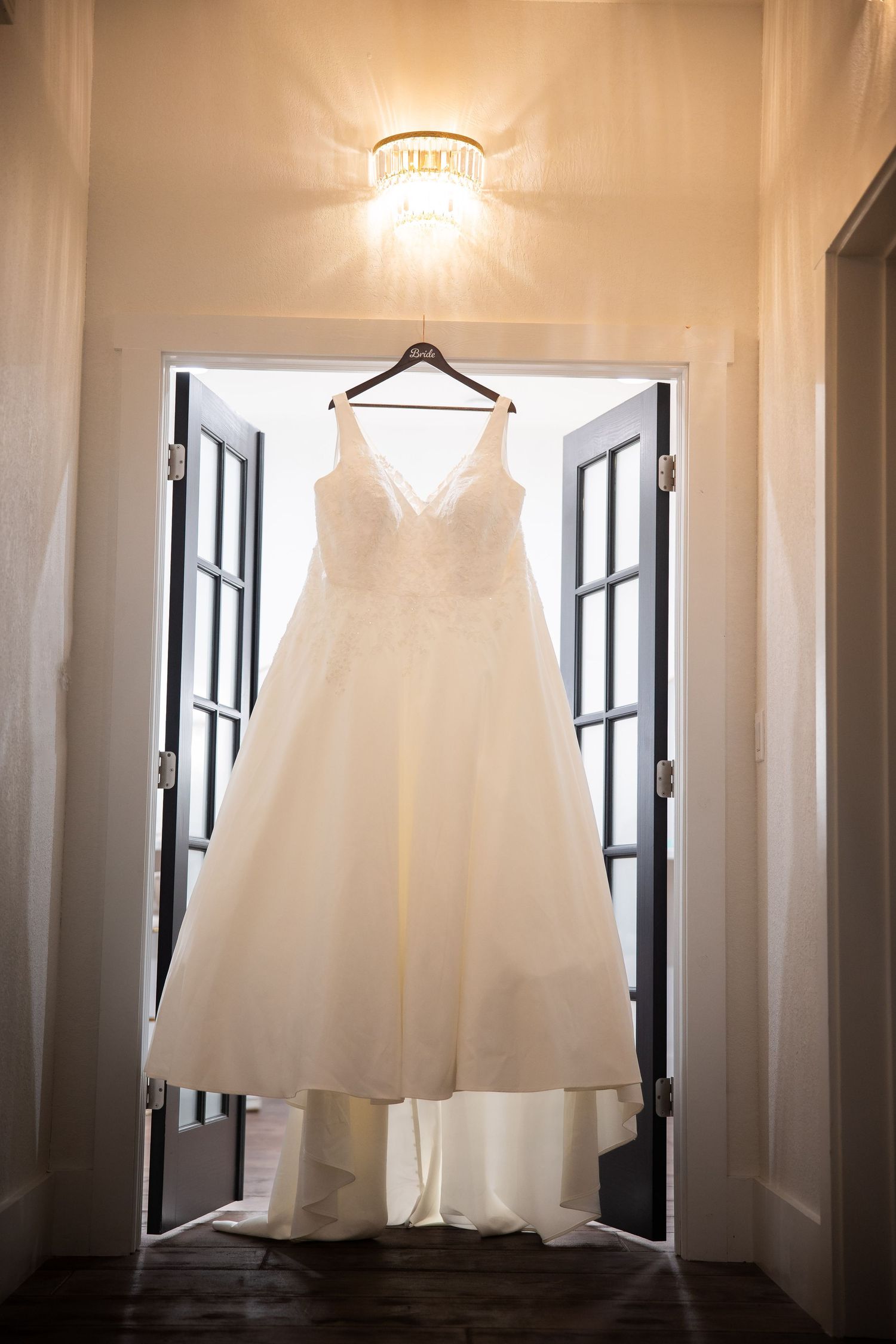 A white wedding dress hangs elegantly in front of a window, creating a romantic silhouette against natural light.