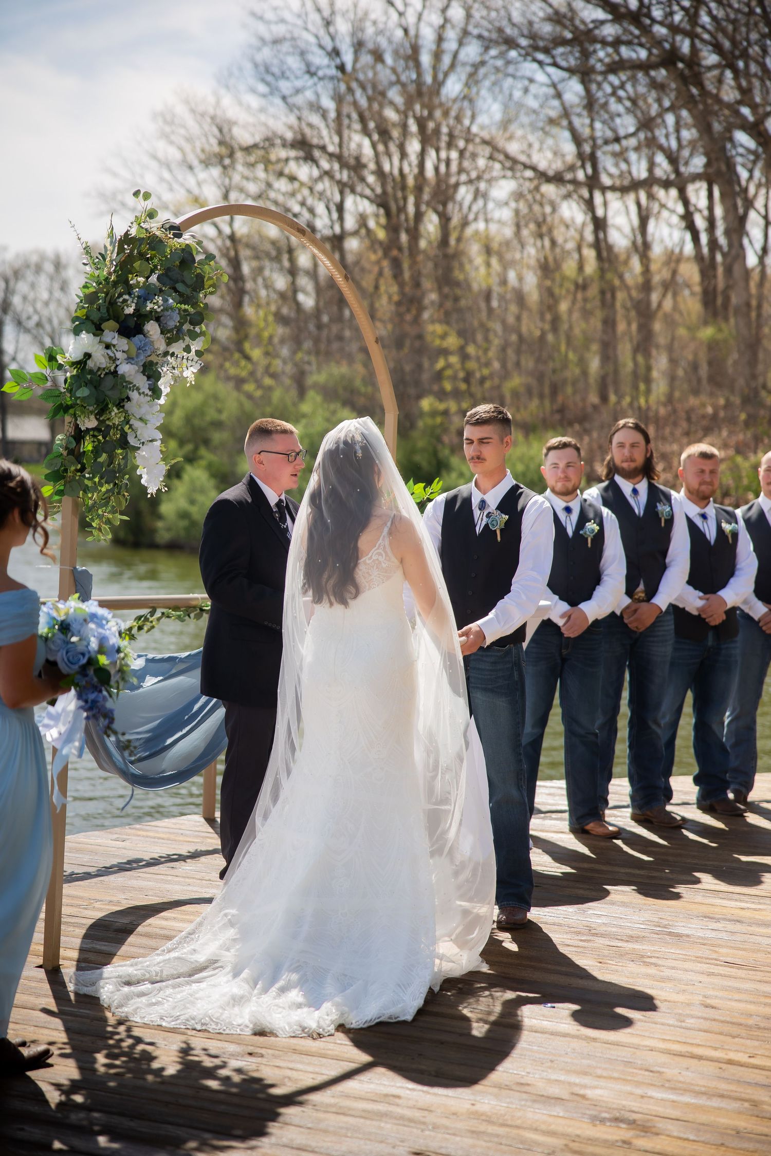 Bride in white gown stands at floral arch altar with groom and groomsmen in vests and jeans on waterfront dock.