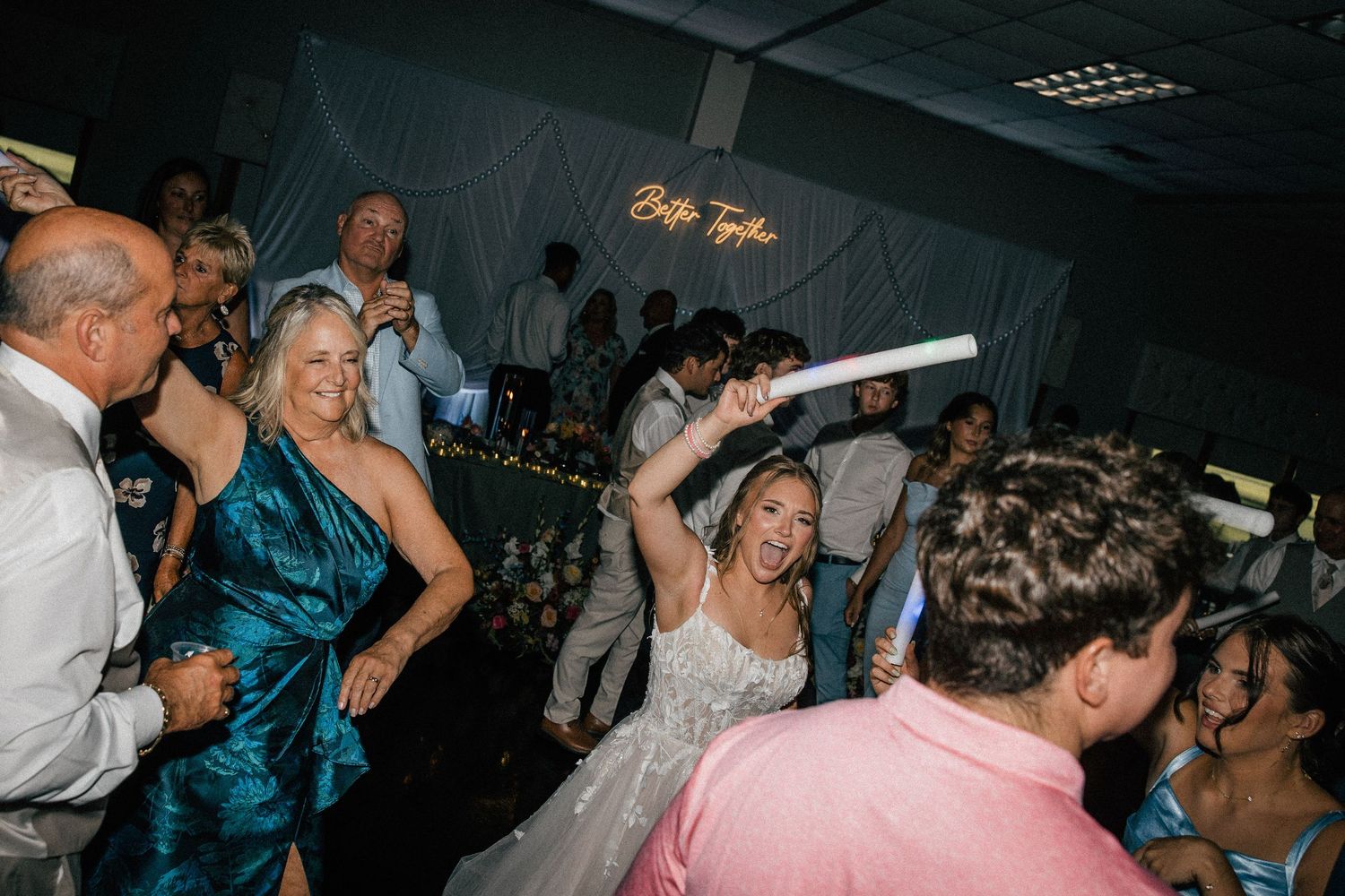 Bride celebrates on dance floor holding glow stick at lively wedding reception with neon sign.