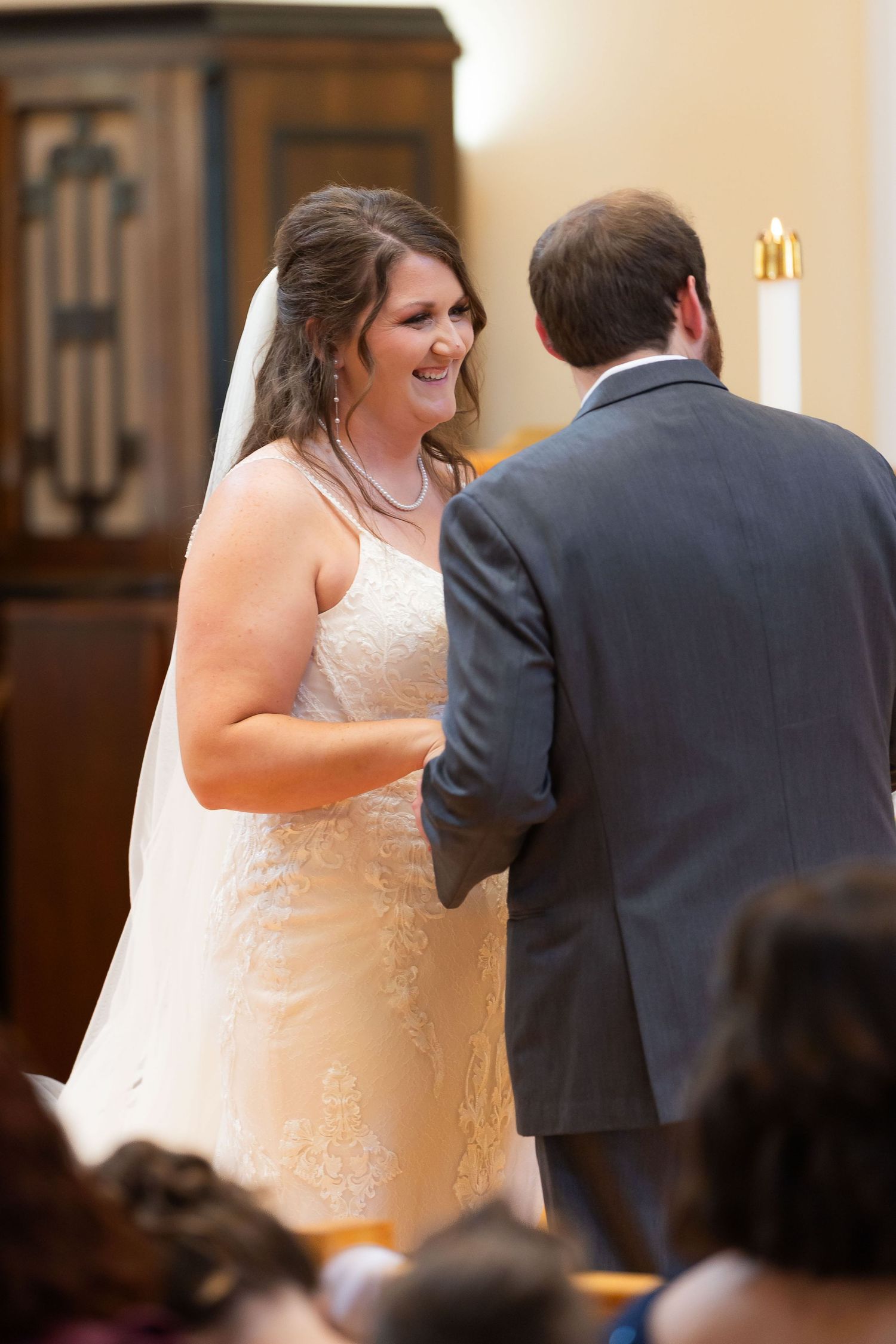 Bride in white lace gown and veil smiles at groom in gray suit during wedding ceremony in church.