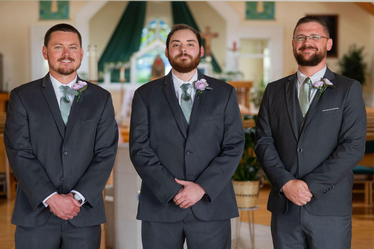Three groomsmen in matching dark suits with mint green ties pose together inside a church before a wedding ceremony.