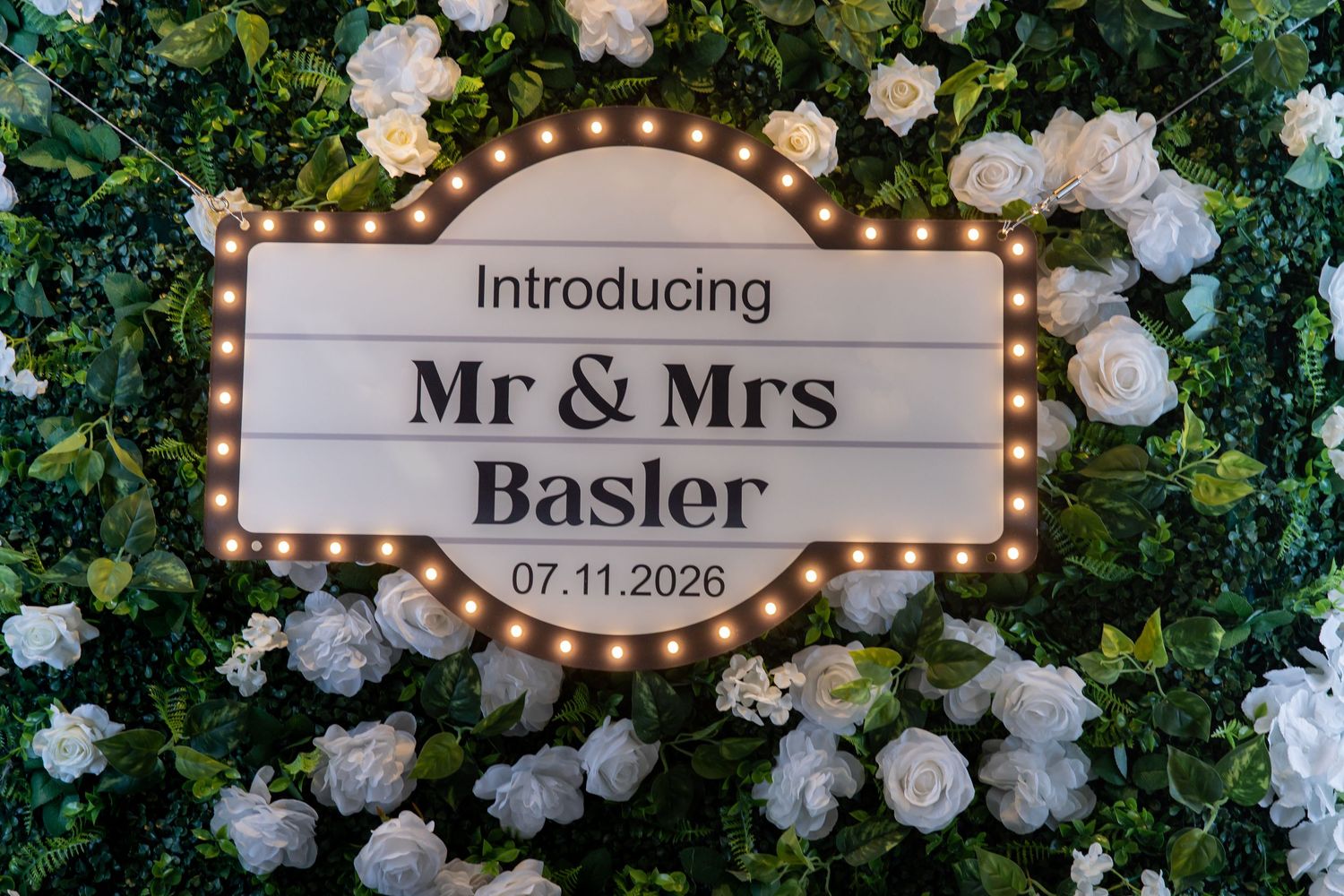 Illuminated marquee sign introducing Mr & Mrs Basler dated 07.11.2026 surrounded by white roses.