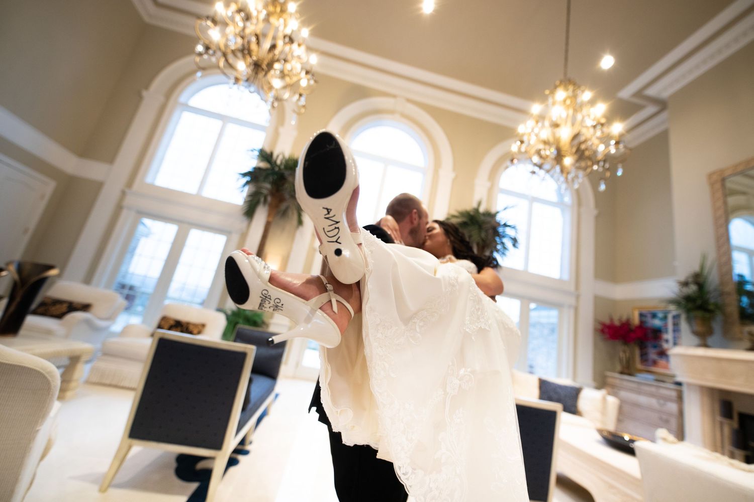 Groom sweeps bride off her feet in elegant chandelier ballroom, her white heels raised high in joyful wedding celebration.
