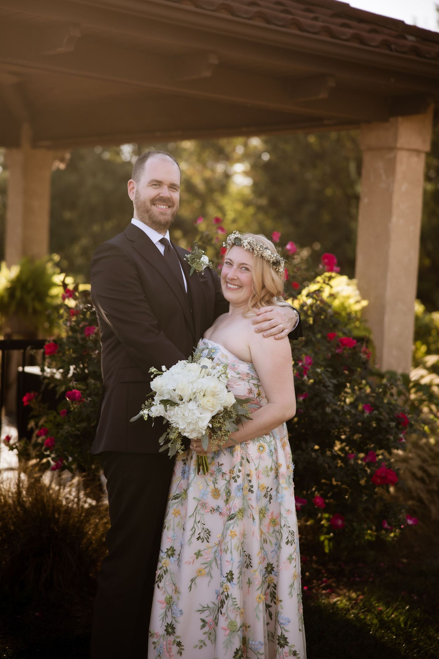 Couple in formal attire poses for romantic wedding photos in a garden setting with blooming roses at sunset.