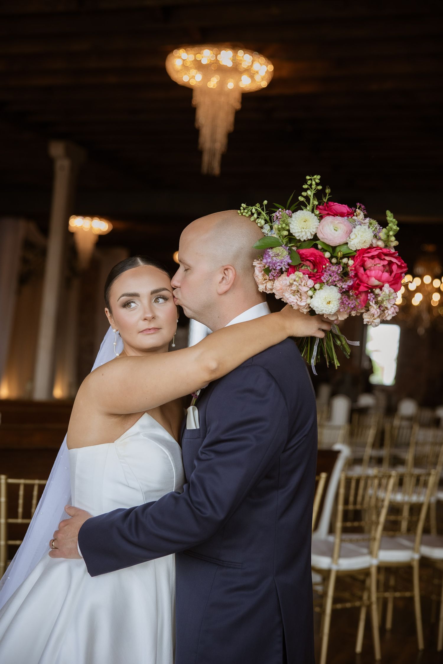 Romantic wedding ceremony moments in an indoor venue with chandeliers and white chairs.