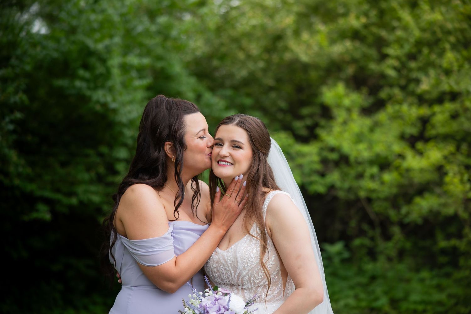 Bridesmaid in lavender dress kisses bride wearing veil and holding floral bouquet in green outdoor setting.