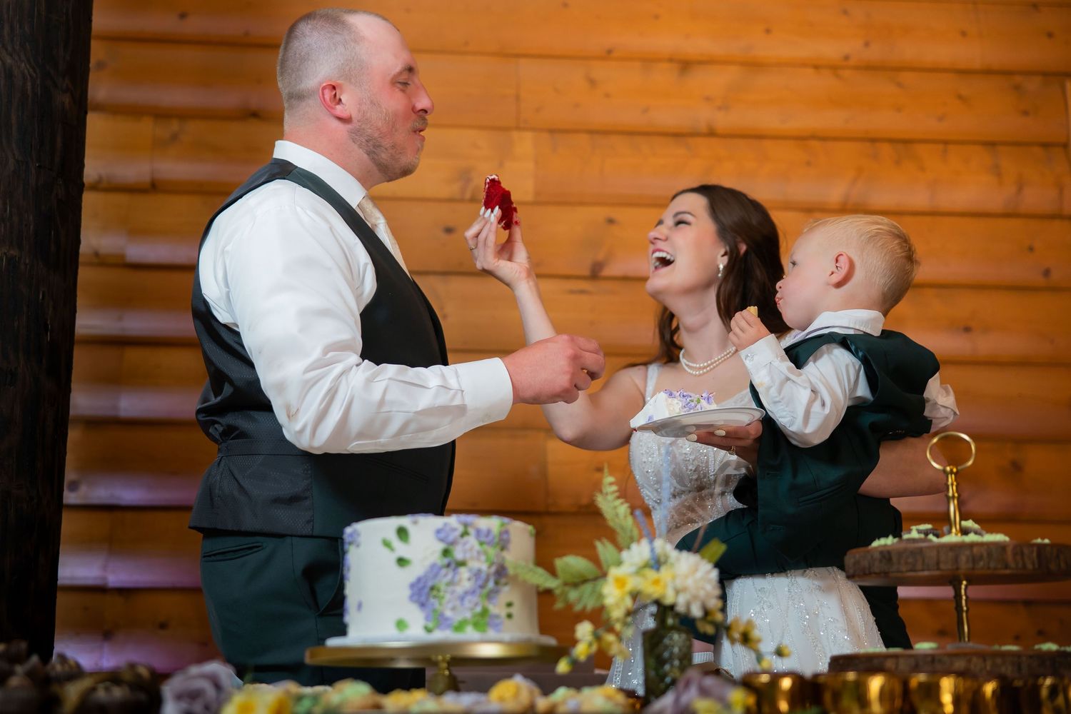 Bride and groom laugh during cake cutting while holding a baby at a rustic log cabin reception.