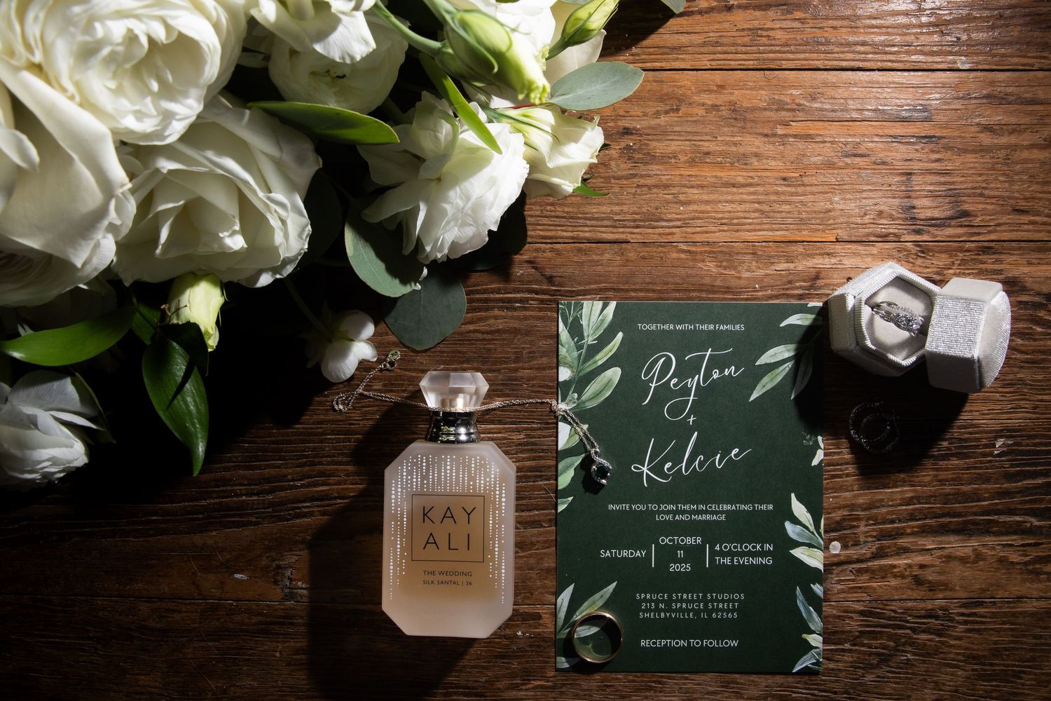Wedding flat lay with white roses, Kayali perfume, green botanical invitation, and diamond rings on rustic wood.