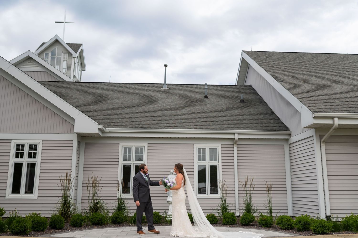Bride and groom share a romantic first look outside a charming gray chapel with a white cross on a cloudy day.
