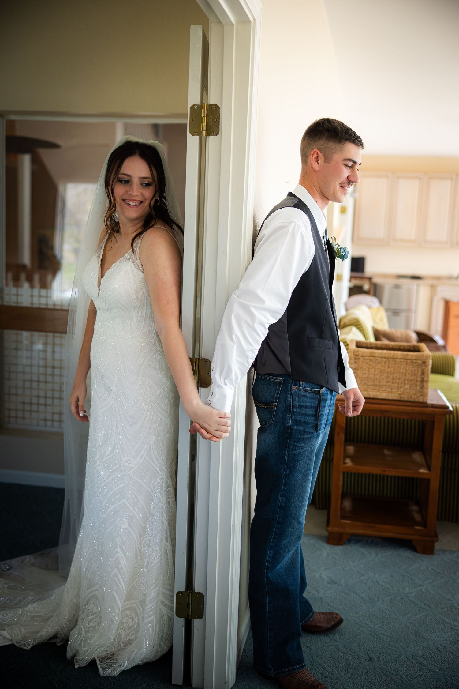 Bride in white lace gown and groom in vest and jeans hold hands around a door in a sweet first look moment.