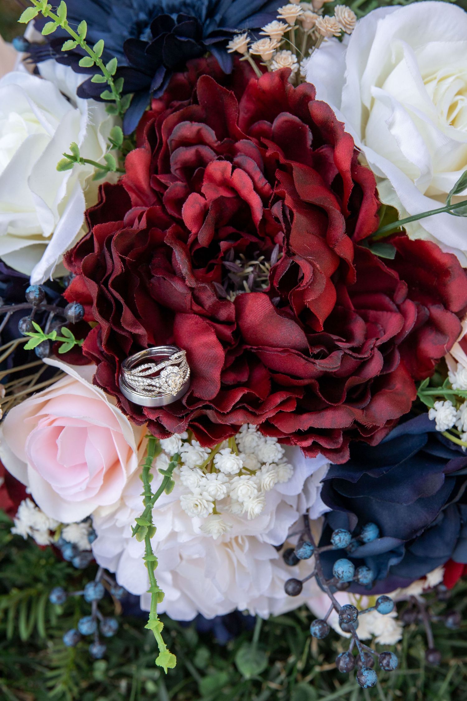 Wedding rings rest on a lush bridal bouquet featuring deep red peonies, blush pink roses, white flowers, and navy blue accents.