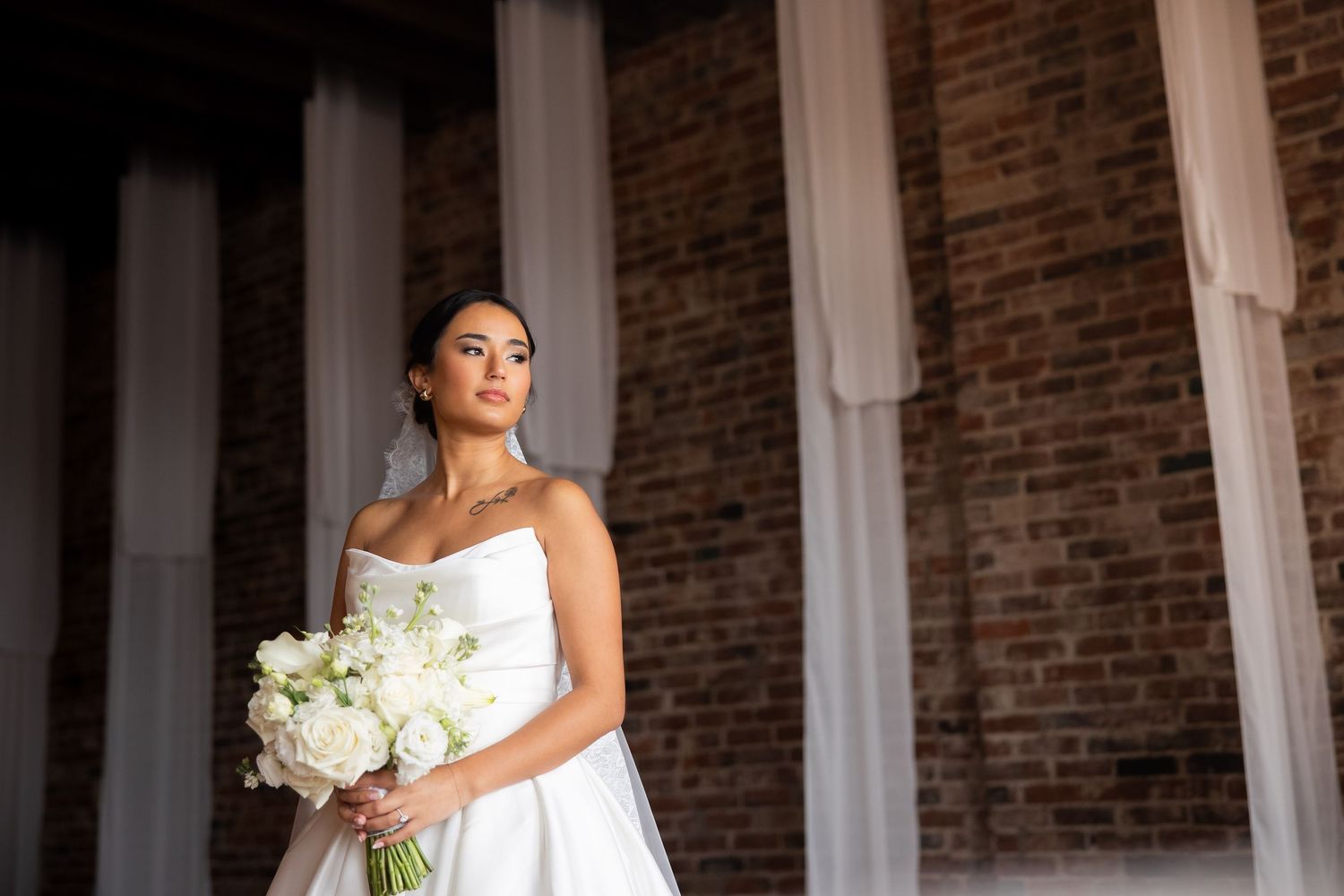 Elegant bride in white strapless gown holds white rose bouquet while posing against rustic brick wall.
