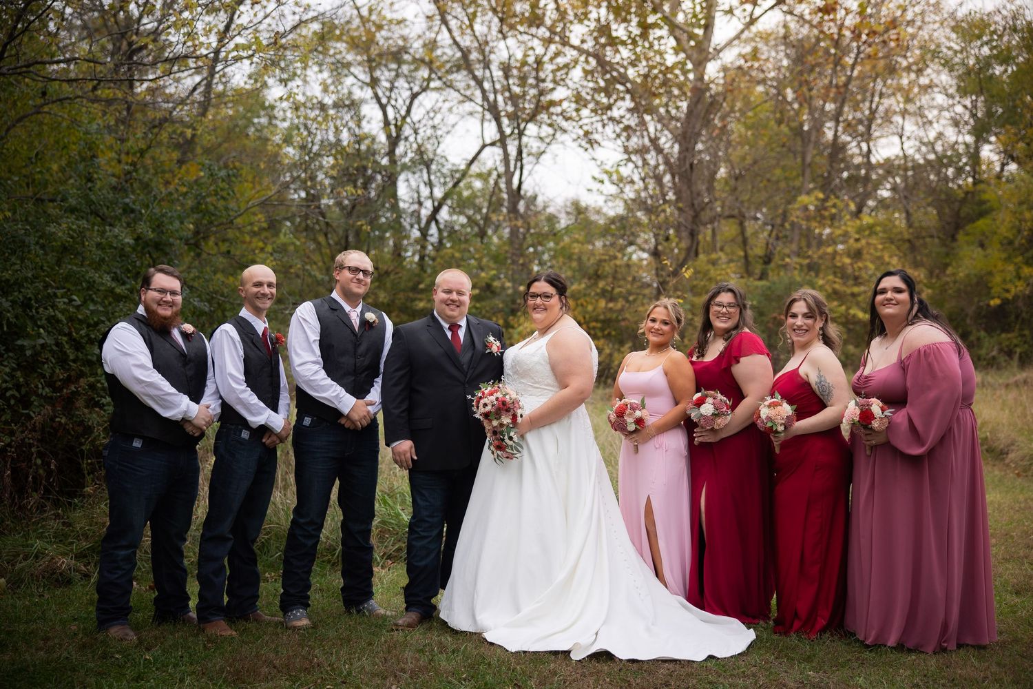 Wedding party in burgundy and mauve attire poses together in front of autumn trees.