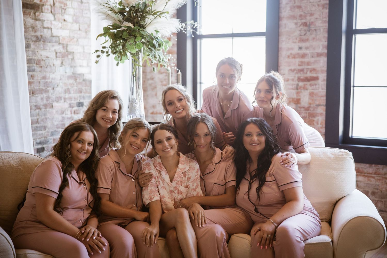Group of bridesmaids in matching pink pajamas smiling together on a couch in a rustic brick-walled room.