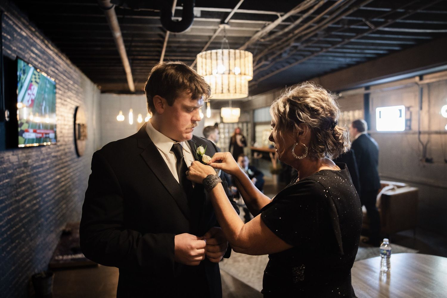 A woman pins a boutonniere on a young man in a black suit at a formal event venue.